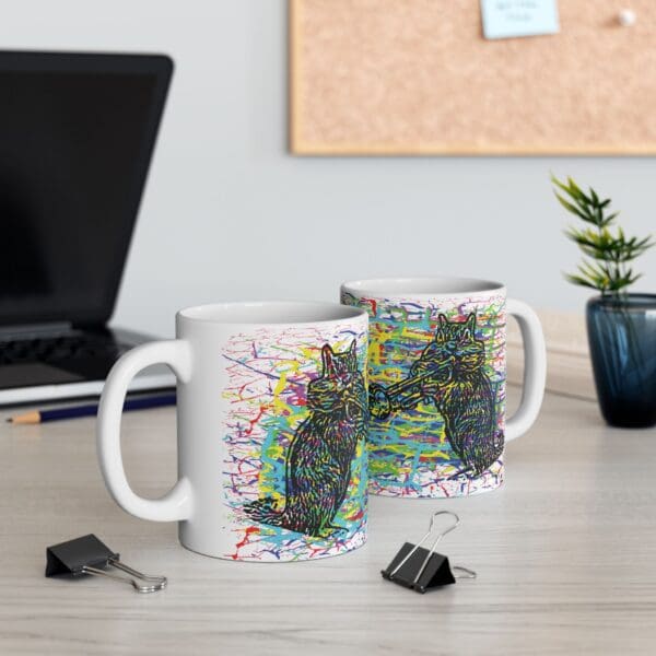 Two Cracked Chipmunk Coffee Mugs featuring colorful cat artwork are placed on a desk. The desk also has a black laptop, two black binder clips, a pen, and a small plant. A corkboard is visible in the background.