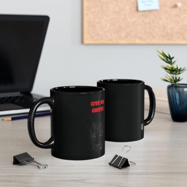 Two black Angry Cat Coffee Mugs, one featuring red text and a graphic, are on a desk next to a laptop, a small potted plant, and binder clips. A corkboard with notes is in the background.