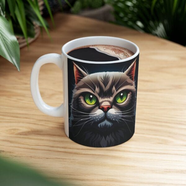 An 11oz Angry Cats Coffee Mug, featuring a detailed illustration of a grumpy-looking cat with green eyes, is filled with a hot beverage and placed on a wooden table. Green leaves are visible in the background.