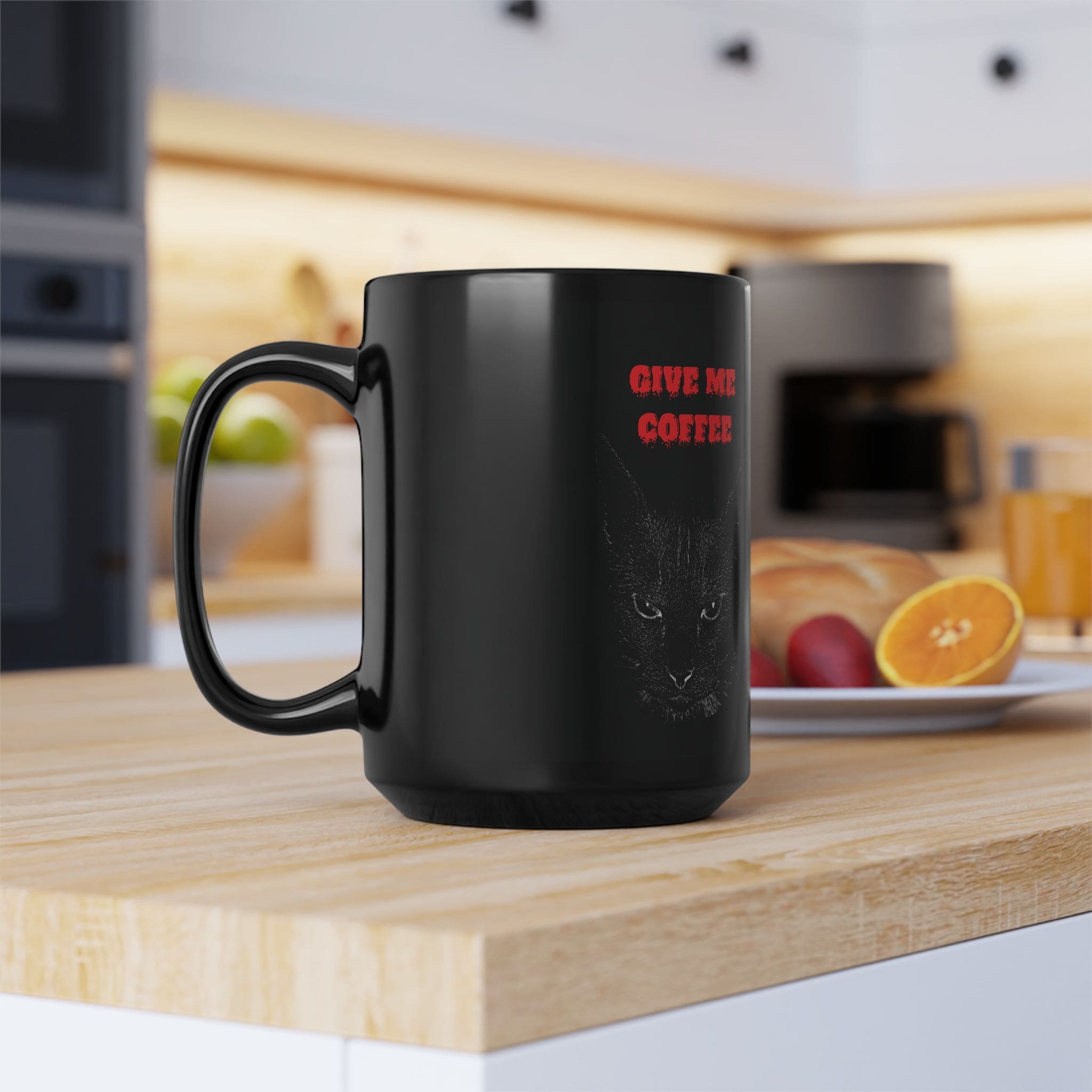 Angry Cat Coffee Mug (available in 11oz and 15oz) with the text "Give Me Coffee" and a cat's face printed on it, placed on a kitchen counter. In the background, there is a plate with fruit and a blurred kitchen setting.