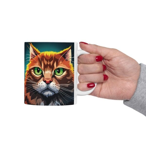 A hand with red nail polish holds the Angry Cats Coffee Mug 11oz, which features a colorful illustration of a cat with green eyes and a vibrant fur pattern.