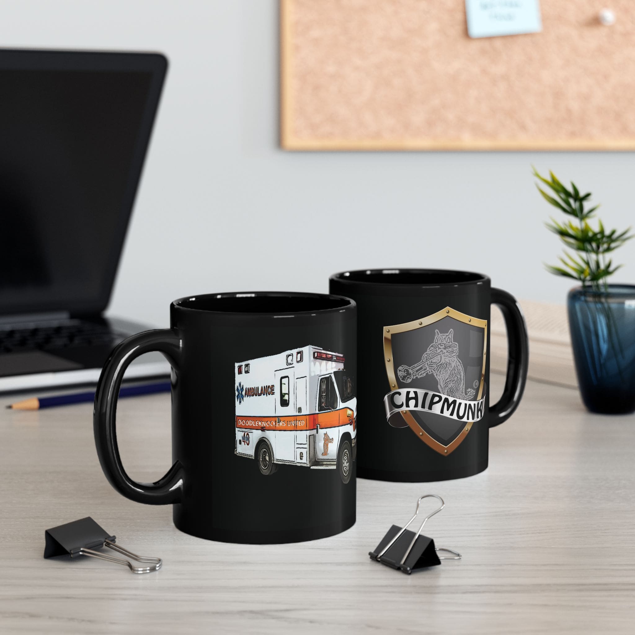 Two black mugs on a desk. The mug on the left is the Ambulance Coffee Mug, featuring an image of an ambulance and the word "Ambulance." The mug on the right has a shield with a chipmunk and the word "Chipmunk." Office items surround them.