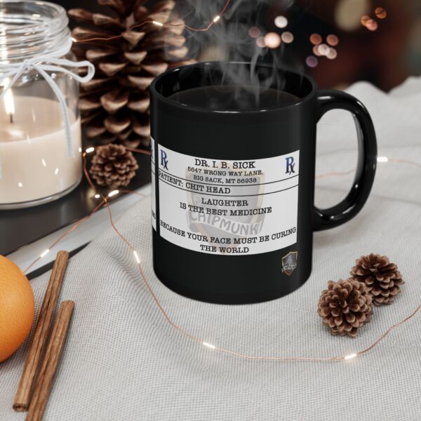 A black "Laughter Is The Best Medicine" coffee mug sits on a table, showcasing its humorous prescription label design. The table is also adorned with cinnamon sticks, pine cones, an orange, and a jar candle.