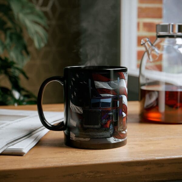 An American Trucker Coffee Cup, featuring a black design with an American flag and a truck motif, emits steam as it sits on a wooden table next to a newspaper and a glass pitcher containing a dark liquid.