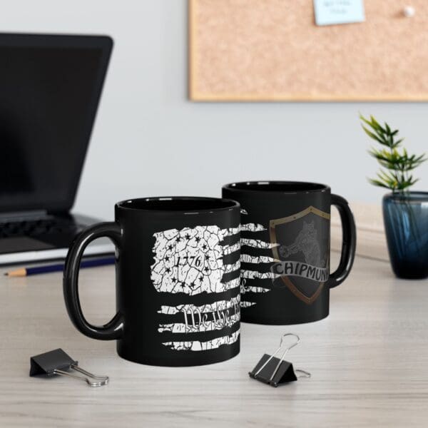 Two American Pride Coffee Mugs, featuring black exteriors with white printed designs, sit on a desk alongside binder clips, a laptop, and a potted plant in the background.