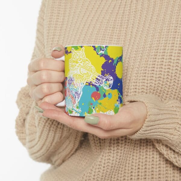 A person holding a white Splash Chipmunk Coffee Mug featuring colorful abstract paint splatters and a trumpet-playing wolf illustration. The person is wearing a beige knitted sweater.