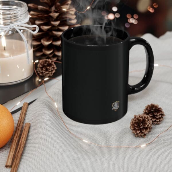 An Angry Cat Coffee Mug sits steaming on a fabric surface surrounded by pine cones, a lit candle, cinnamon sticks, an orange, and a string of lights in the background.