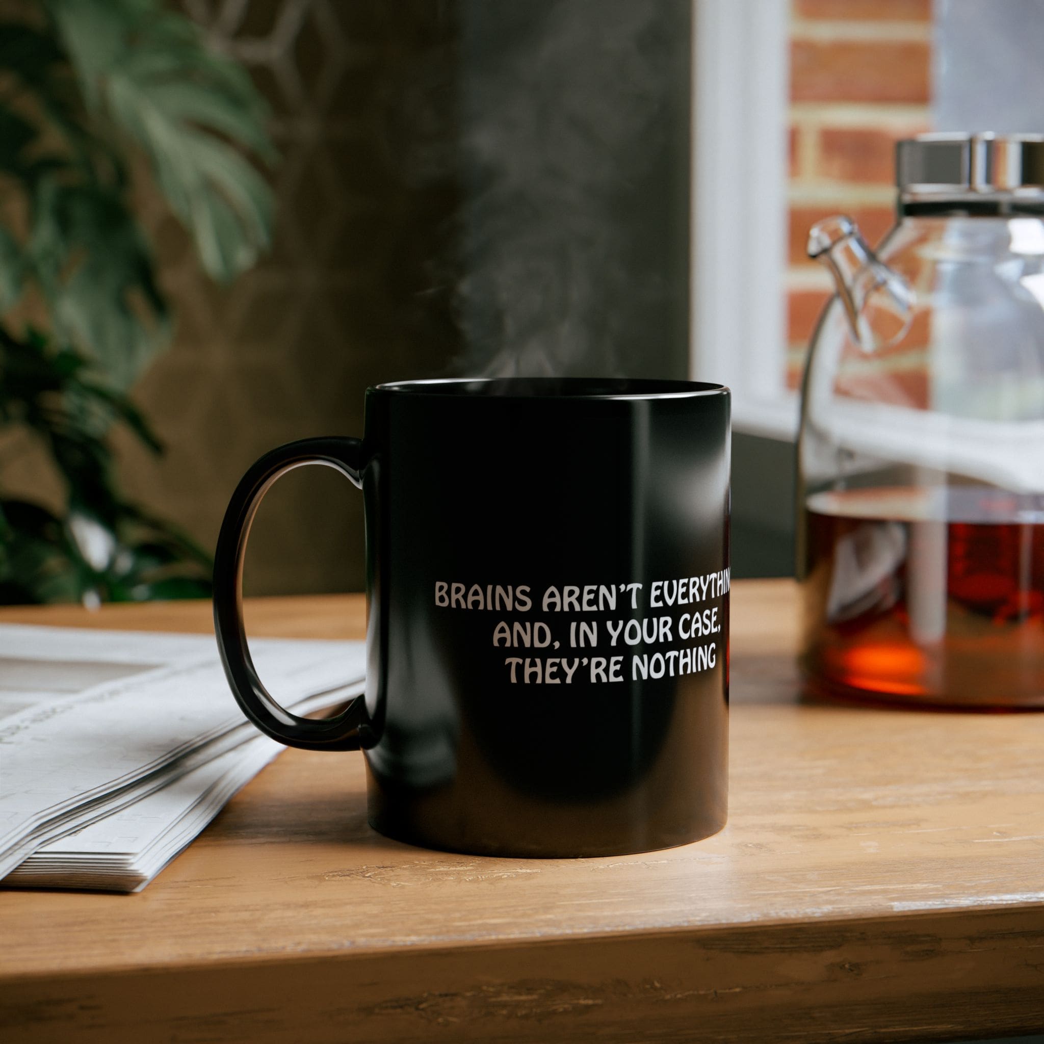 A black "Brains Aren't Everything Coffee Mug" on a wooden table with the text "BRAINS AREN'T EVERYTHING AND, IN YOUR CASE, THEY'RE NOTHING" printed on it, next to a newspaper and a glass container with liquid.