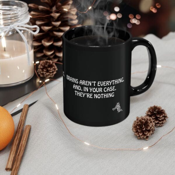 A Brains Aren't Everything Coffee Mug, featuring the phrase "Brains aren't everything. And, in your case, they're nothing," sits on a table surrounded by pinecones, cinnamon sticks, and a candle.