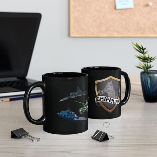 Two black mugs on a desk, one featuring illustrations of military vehicles belonging to the "Land, Sea, and Air Coffee Mug" collection and the other showcasing a "Chipmunk" shield logo. Also on the desk are a laptop, plant, binder clips, and a corkboard in the background.