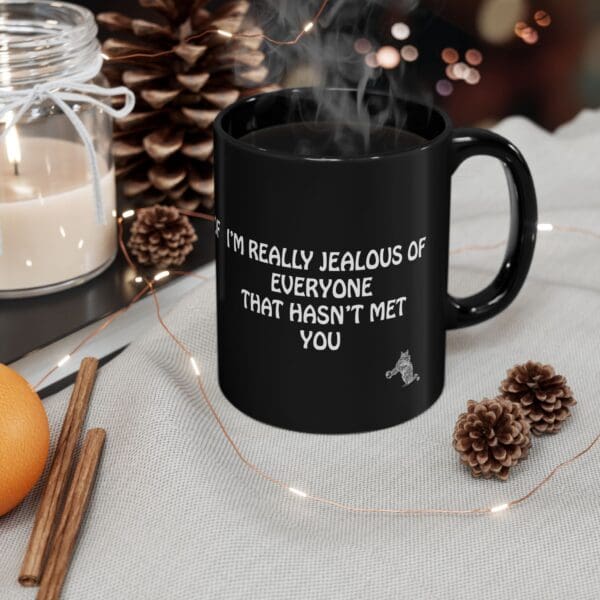 A black Jealous Coffee Mug with the phrase "I'M REALLY JEALOUS OF EVERYONE THAT HASN'T MET YOU" sits next to a lit candle, pinecones, and cinnamon sticks.