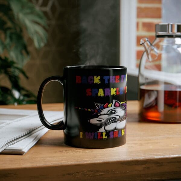 A Unicorn Coffee Mug - Back the F*** Up, Sparkle Tits! featuring a cartoon unicorn and colorful text declaring, "Back the F**k up Sparkle T**ts, I will shank you," sits on a wooden table next to a glass pitcher and some newspapers.