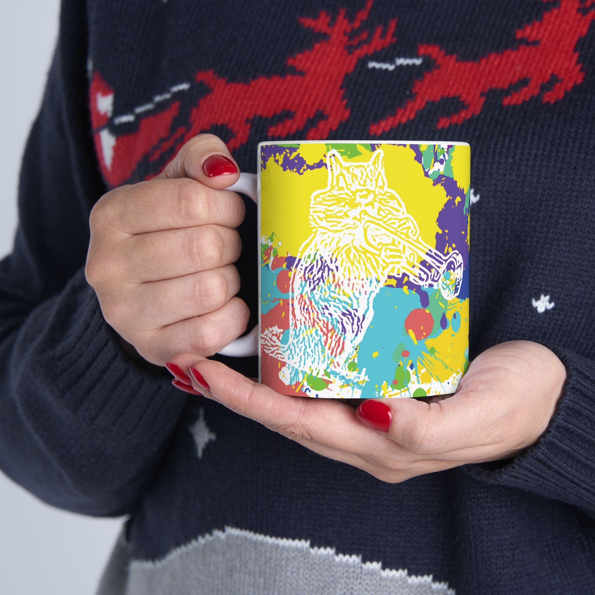 A person holding the Splash Chipmunk Coffee Mug, which features a vibrant pattern with a wolf illustration and abstract splashes of paint. The person is dressed in a dark sweater adorned with a red and white design.