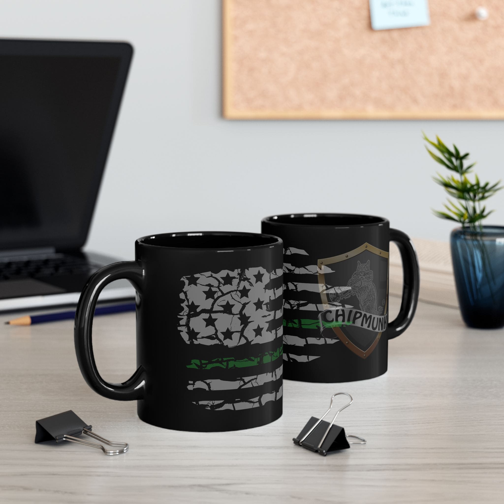 Two black Military Support Coffee Mugs featuring faded American flag designs and the text "CHIPMUNK" on one are situated on a desk, accompanied by a laptop, binder clips, and a small plant in the background.