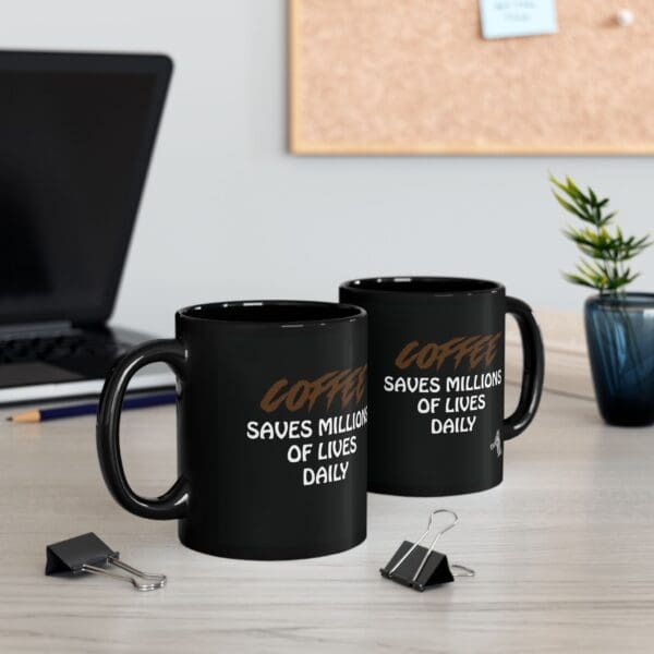 Two "Coffee Saves Lives" black coffee mugs sit on a desk next to binder clips, a plant, a laptop, and a corkboard in the background.