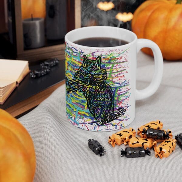 A Cracked Chipmunk Coffee Mug featuring a sketched cartoon cat is filled with a steaming beverage, surrounded by Halloween-themed candies and pumpkins on a grey cloth.