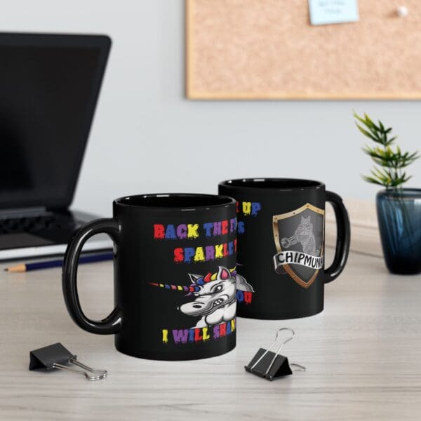 On the desk are two black coffee mugs; one showcasing the Unicorn Coffee Mug - Back the F*** Up, Sparkle Tits! with a vibrant unicorn design and text, and the other presenting a shield emblazoned with "CHIPMUNK". Additionally, a laptop, plant, and binder clips can be seen on the desk.