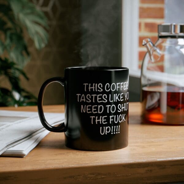 A Shut Up Coffee Mug with a humorous and explicit message printed on it, sitting on a wooden table beside a newspaper and a glass coffee pot.