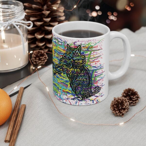 A Cracked Chipmunk Coffee Mug, with a colorful illustration of a hand-drawn cat, sits on a table next to a candle, pine cones, cinnamon sticks, and a piece of citrus fruit.