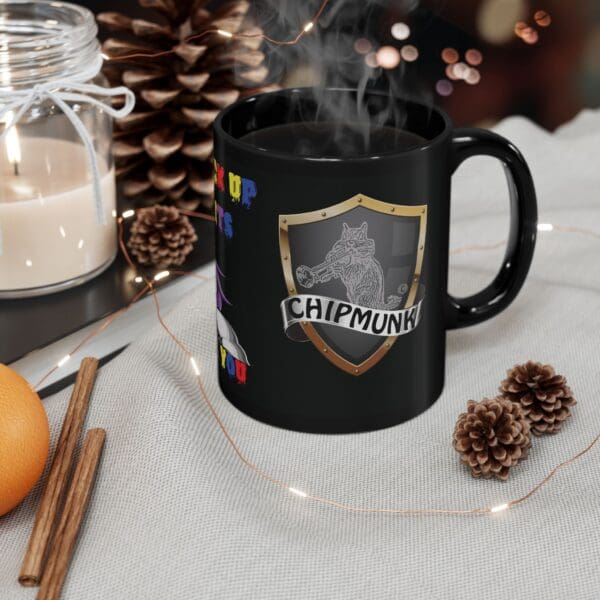 A black "Unicorn Coffee Mug - Back the F*** Up, Sparkle Tits!" with a wolf graphic. It's placed on a table next to a lit candle, pinecones, cinnamon sticks, an orange, and a string of lights.