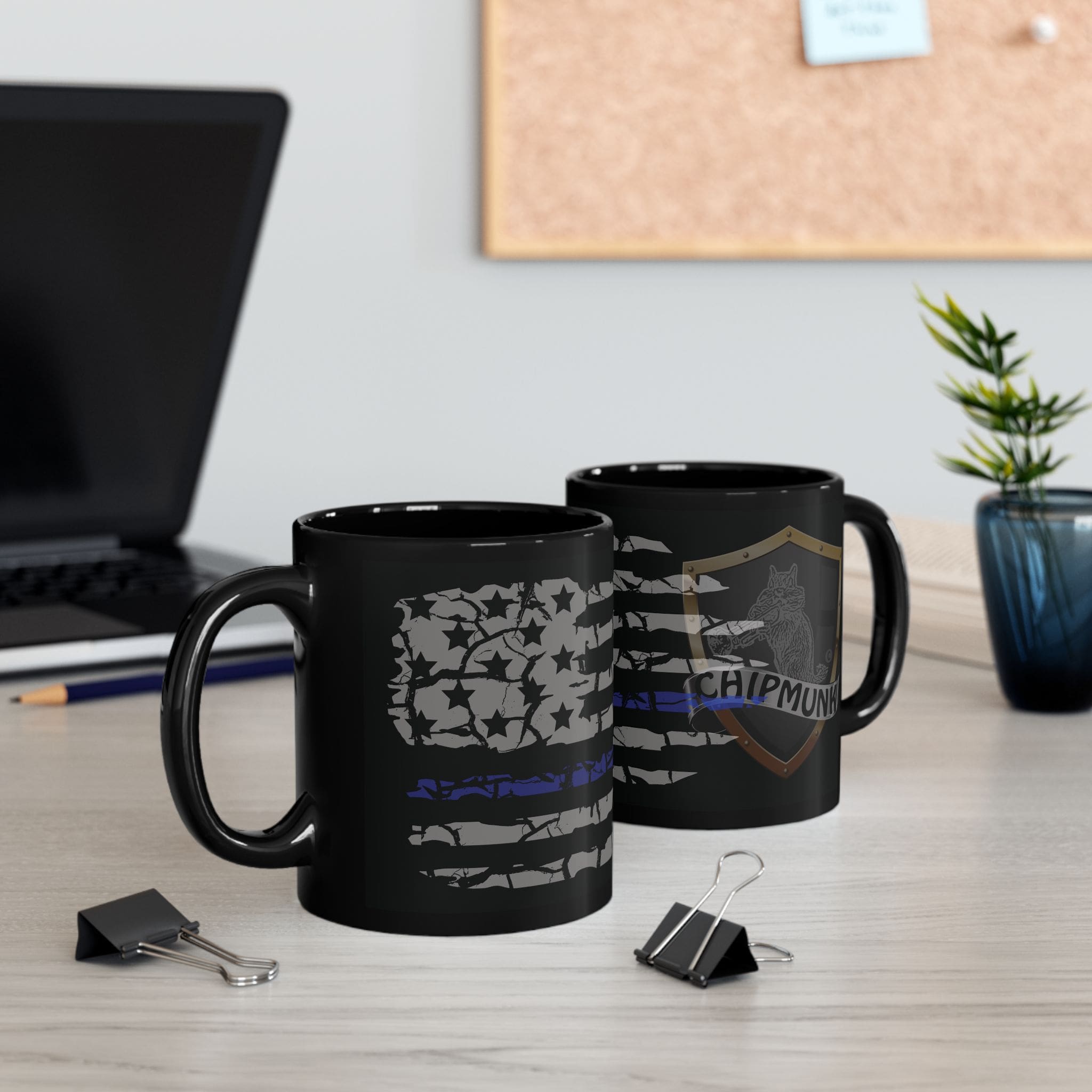 Two Police Support Coffee Mugs with graphic designs sit on a desk near a laptop, two binder clips, a small plant, and a corkboard on the wall in the background.