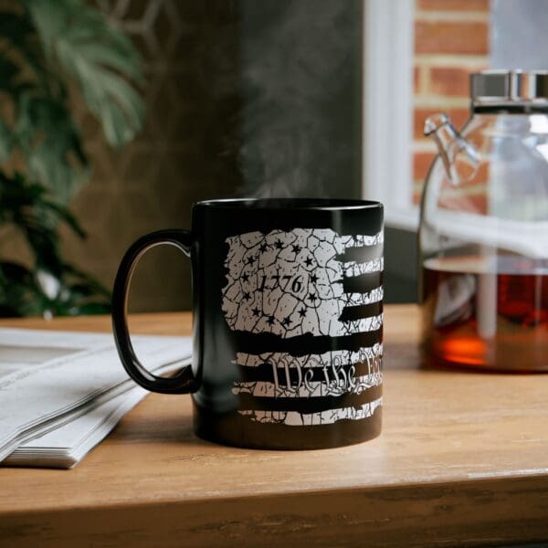 An American Pride Coffee Mug, featuring a "1776" and American flag design, sitting steaming on a wooden table beside a newspaper, with a glass coffee carafe in the background.