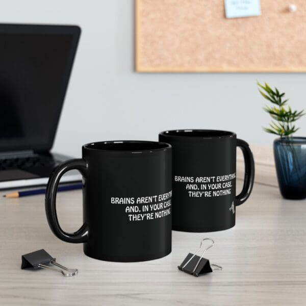 Two "Brains Aren't Everything" coffee mugs on a desk with a witty quote, two binder clips, a potted plant, a laptop, and a corkboard in the background.