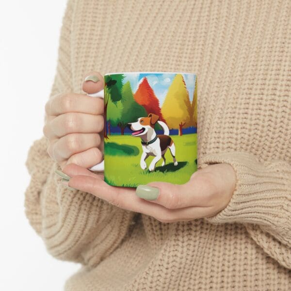 A person wearing a beige sweater holds the Playful Dog Coffee Mug 11oz, which features an illustration of a dog standing on grass with trees in the background.