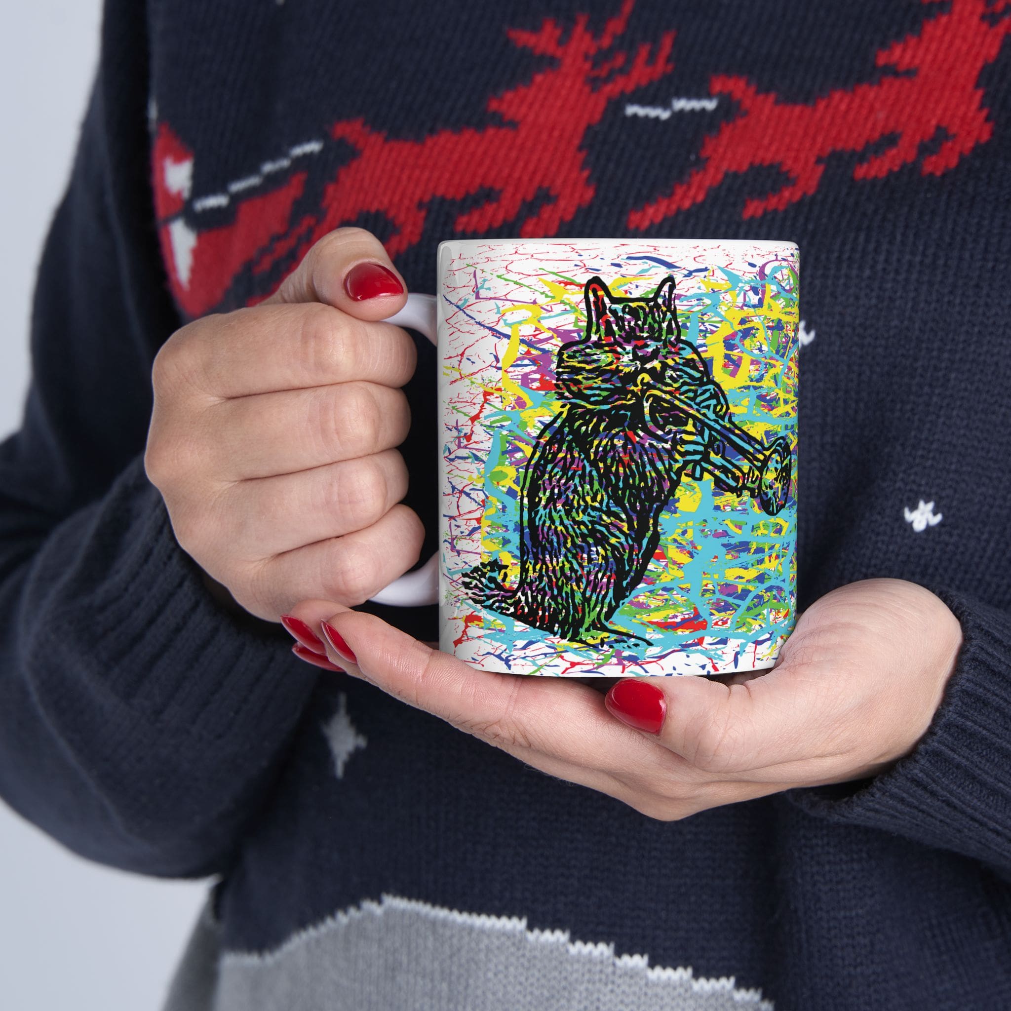 A person wearing a sweater with red reindeer designs holds a colorful Cracked Chipmunk Coffee Mug featuring an abstract, vibrant cat illustration with red, blue, and yellow splashes.