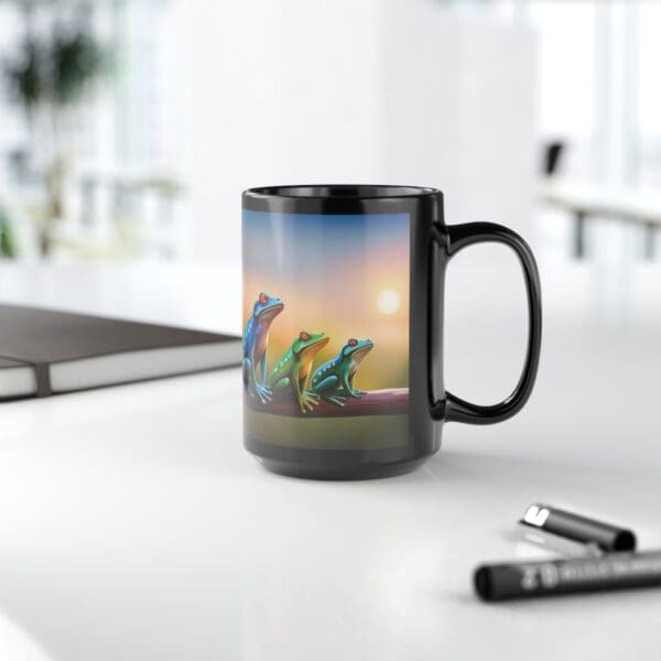 A Colorful Frogs Coffee Mug (available in 11oz and 15oz) featuring an illustration of three vibrant frogs sits on a white desk, accompanied by two markers and an open notebook. A bright window and office space can be seen in the background.