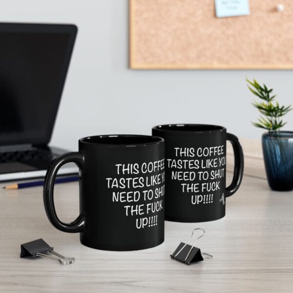 Two "Shut Up Coffee Mug" black coffee mugs on a desk display an explicit humorous message. Nearby are a laptop, binder clips, a small plant, and a corkboard in the background.