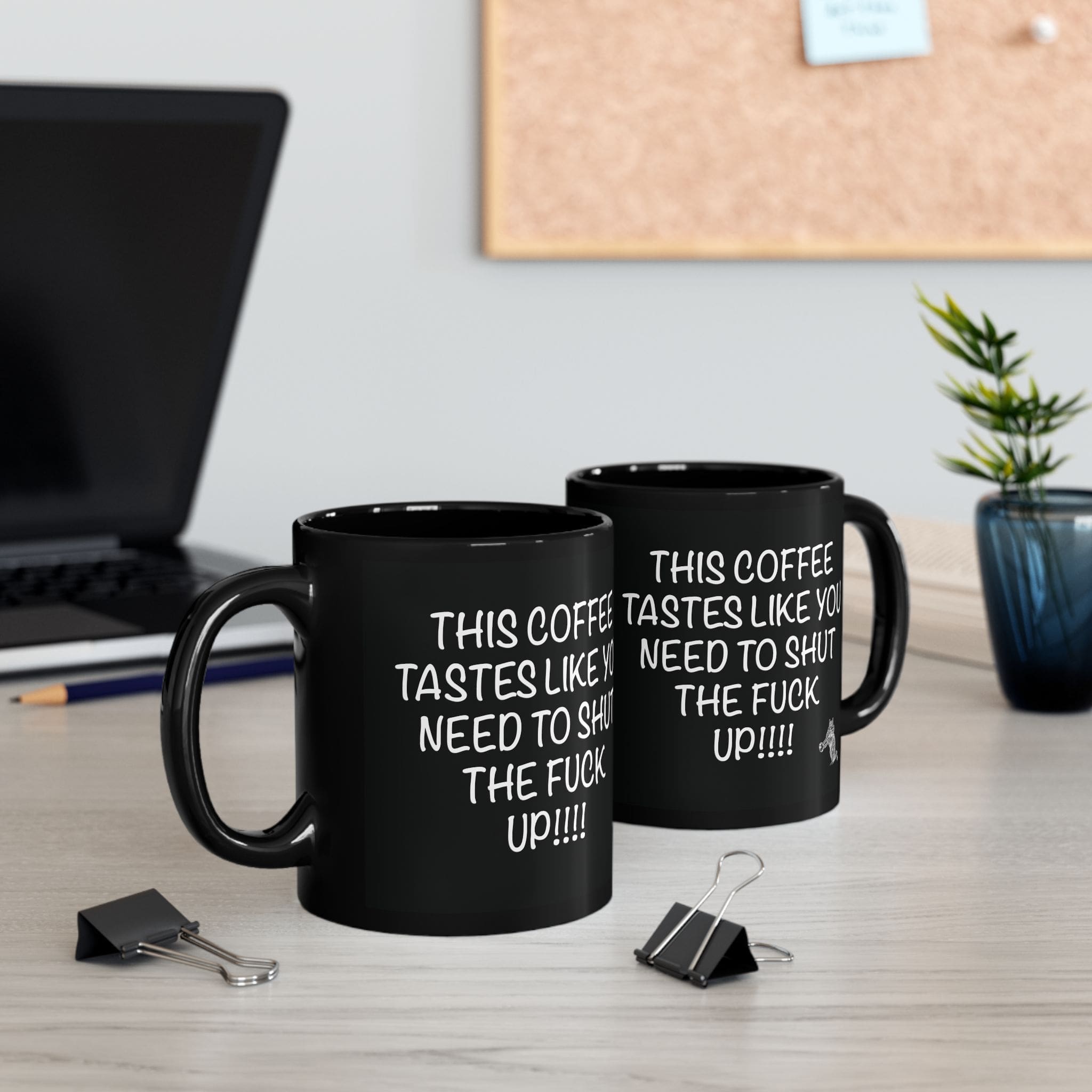 Two "Shut Up Coffee Mug" black coffee mugs on a desk display an explicit humorous message. Nearby are a laptop, binder clips, a small plant, and a corkboard in the background.