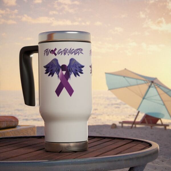 A F*ck Cancer Stainless Steel Travel Mug with Handle, 14oz featuring a purple ribbon with wings design is placed on a table. In the background, there's a beach scene with a sun umbrella and the ocean.