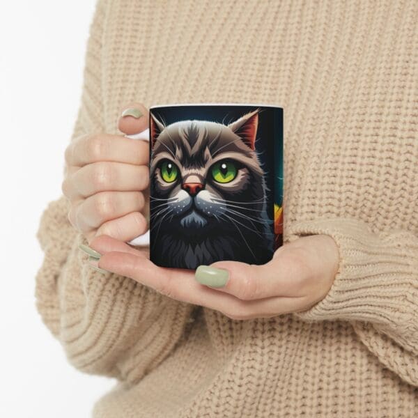 Person in a beige sweater holding an Angry Cats Coffee Mug 11oz featuring an illustrated cat with green eyes and a gray and black fur pattern.