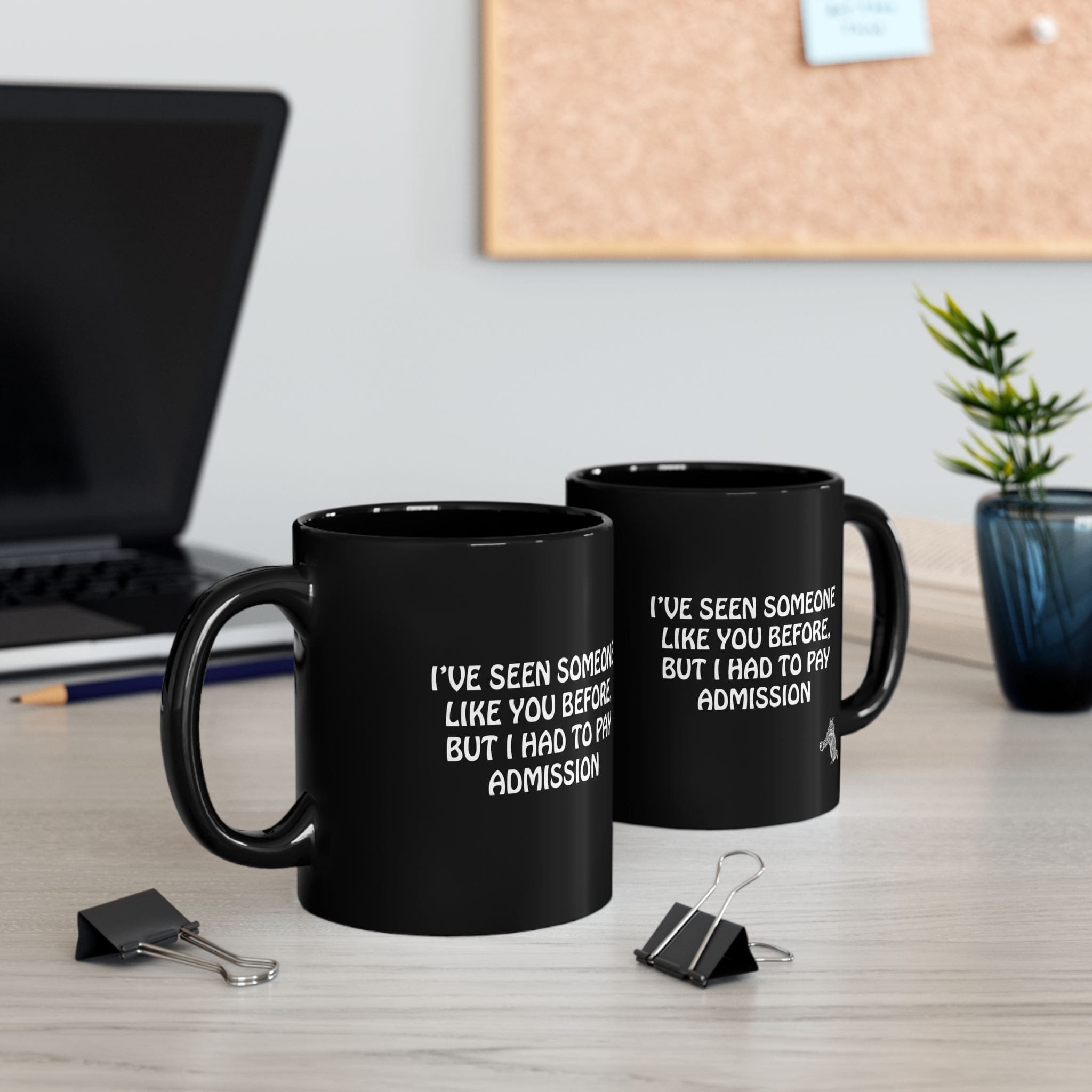 Two Admission Coffee Mugs, featuring black exteriors with white text that reads: "I'VE SEEN SOMEONE LIKE YOU BEFORE BUT I HAD TO PAY ADMISSION," placed on a desk alongside a laptop, paper clips, and a small plant.