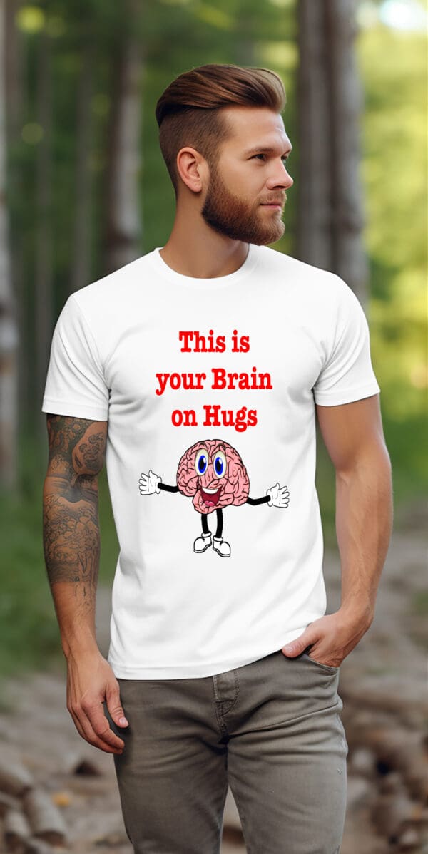 A man with a beard and tattoos is wearing the 'This is Your Brain on Hugs' T-Shirt, which features an illustration of a happy brain with arms and legs. He is standing outdoors on a path.