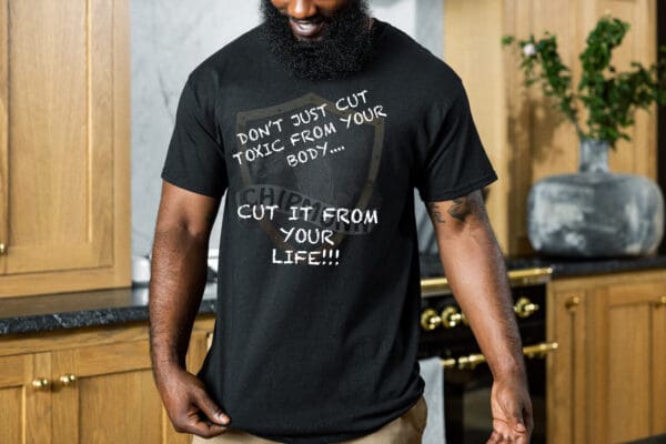 A person in a kitchen wearing the "Don't Just Cut Toxic from Your Body, Cut It from Your Life!" T-Shirt, smiling and looking down.