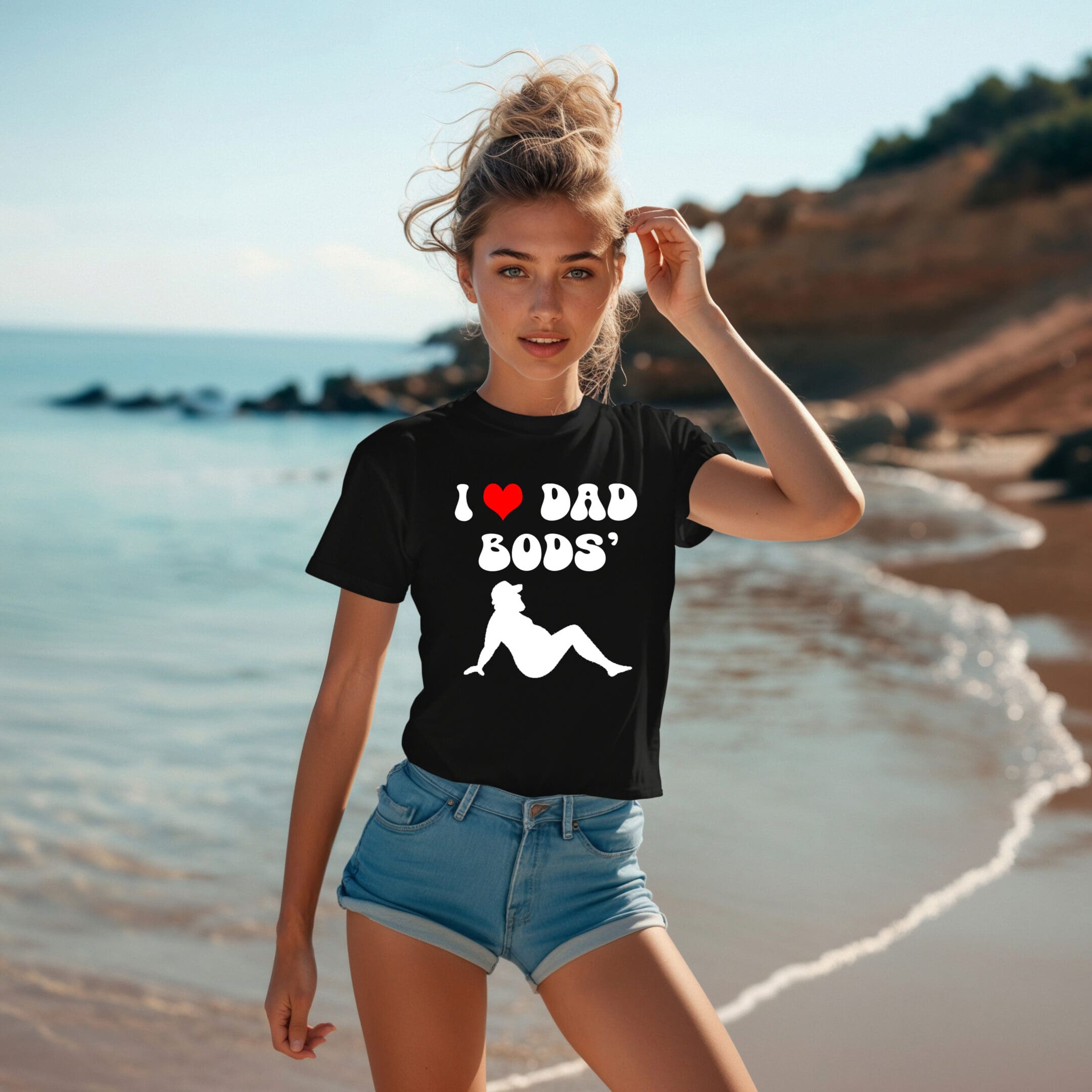 A person stands on a beach wearing an I ❤️ Dad Bods T-Shirt in black, featuring an illustration of a person lounging. The individual, who has blonde hair, is also dressed in denim shorts.