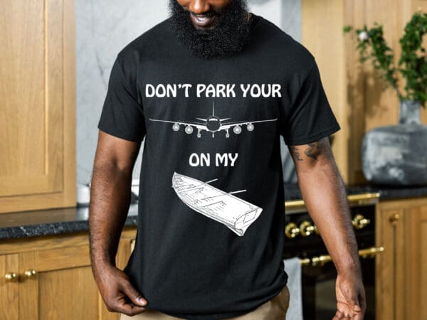 Person wearing a black T-shirt with an illustration of an airplane landing on a dinghy and text that reads, "Don't Park Your Plane on My Dinghy." Kitchen background visible.