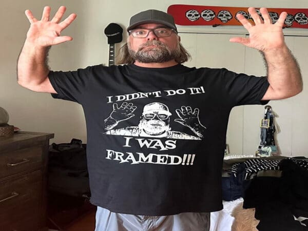 A man with a beard and glasses is raising his hands, wearing a black T-shirt that says, "I didn't do it! I was framed!!!". He stands in a room with various objects in the background.