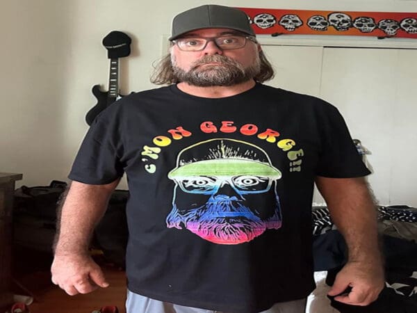 A man with a beard and glasses wears a dark-colored hat and a black T-shirt featuring a colorful graphic and the text "C'MON GEORGE!" in a room with a wall-mounted guitar and skull decorations.