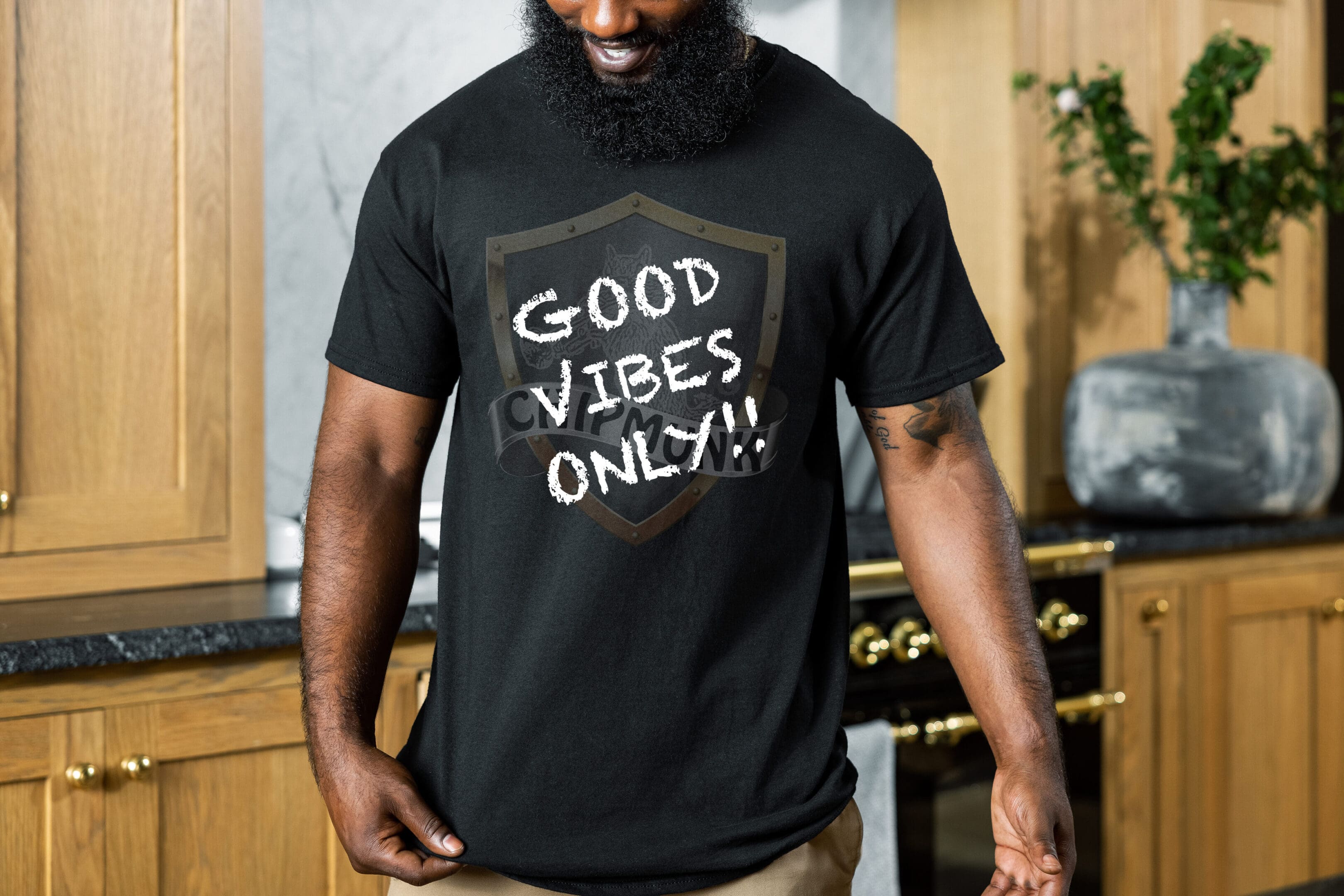 A man in a kitchen wearing the "Good Vibes Only Shirt," which features "Good Vibes Only!" in white chalk-like letters. He is looking down and smiling, with kitchen cabinets and a potted plant visible in the background.