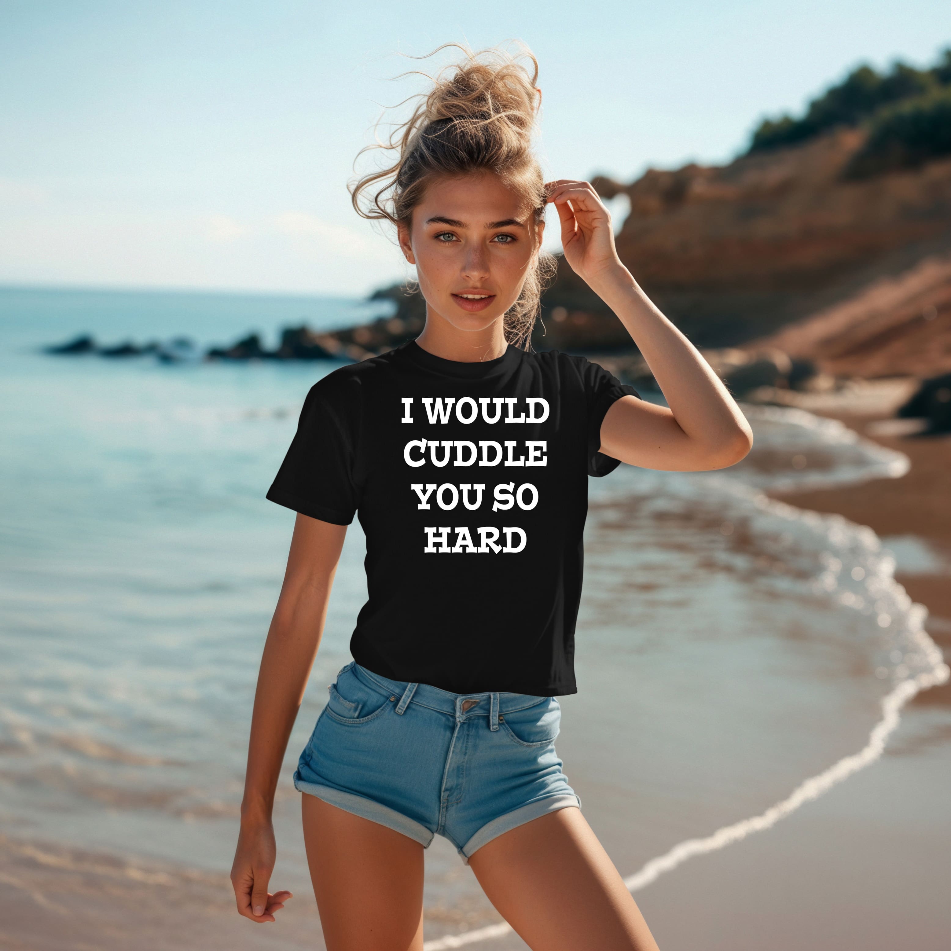 A woman stands on a beach wearing an "I Would Cuddle You So Hard" T-Shirt. She has one hand in her hair and another by her side.