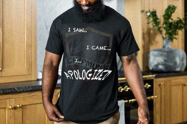 A person with a beard wearing the "I Saw T-Shirt," which is black and reads "I saw... I came... I... APOLOGIZED," stands in a kitchen with wooden cabinets and a black stove in the background.