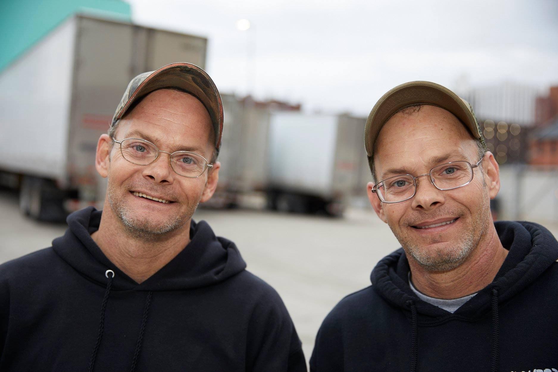 Two men wearing hats and glasses smiling for the camera.