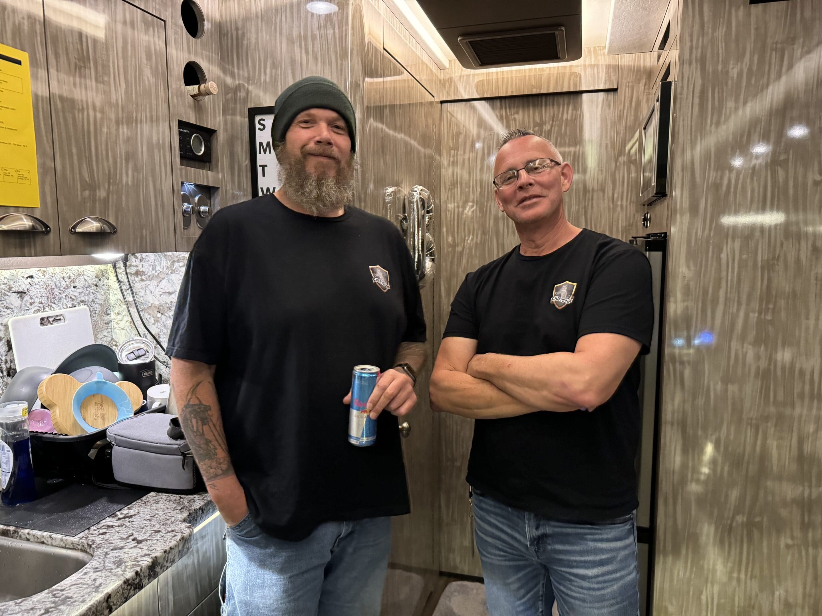 Two men standing in a modern kitchen area. One holds a beverage can and both are wearing black t-shirts with a small logo on the chest. The kitchen counter has various dishes and kitchen items.