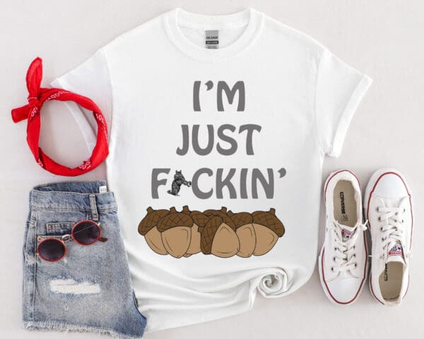 The "I'm Nuts T-Shirt" is a white top featuring the text "I'M JUST F*CKIN'" along with an image of acorns. It is styled with denim shorts, a red bandana, white sneakers, and sunglasses.