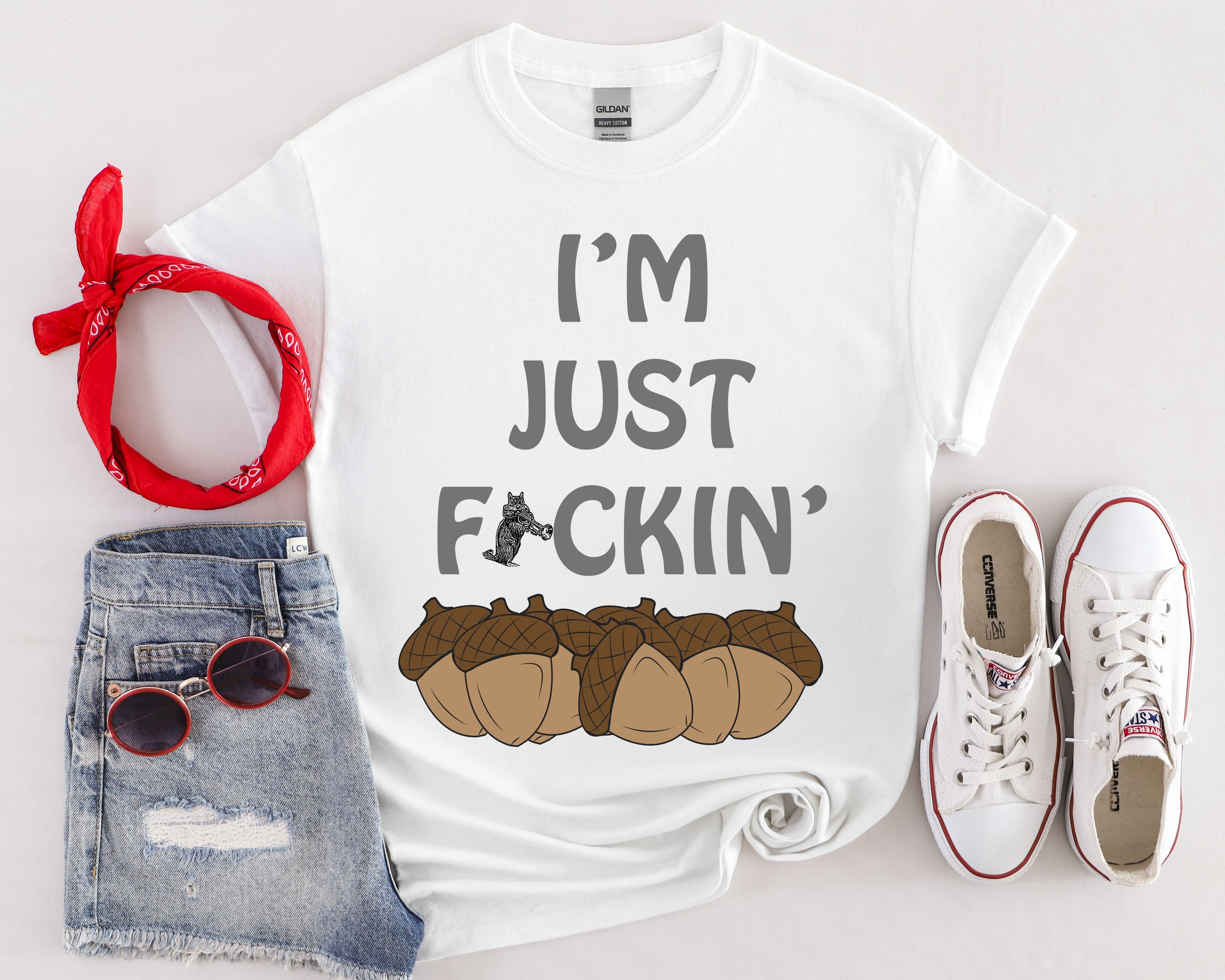 The "I'm Nuts T-Shirt" is a white top featuring the text "I'M JUST F*CKIN'" along with an image of acorns. It is styled with denim shorts, a red bandana, white sneakers, and sunglasses.