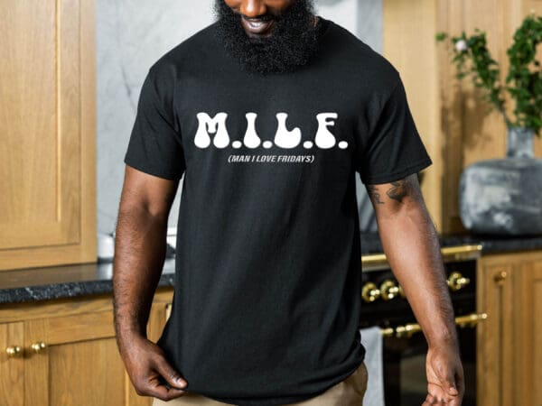 A person wearing a M.I.L.F. (Man I Love Fridays) T-Shirt stands in a kitchen, showcasing the black t-shirt with white text.