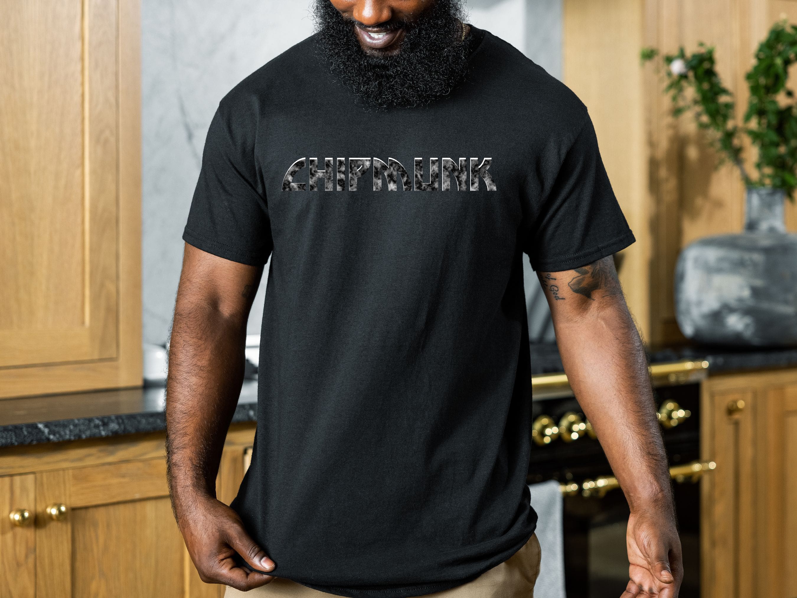 A man with a beard wearing a black Chipmunk Rock T-Shirt #6 stands in a kitchen with wooden cabinets and a black countertop.